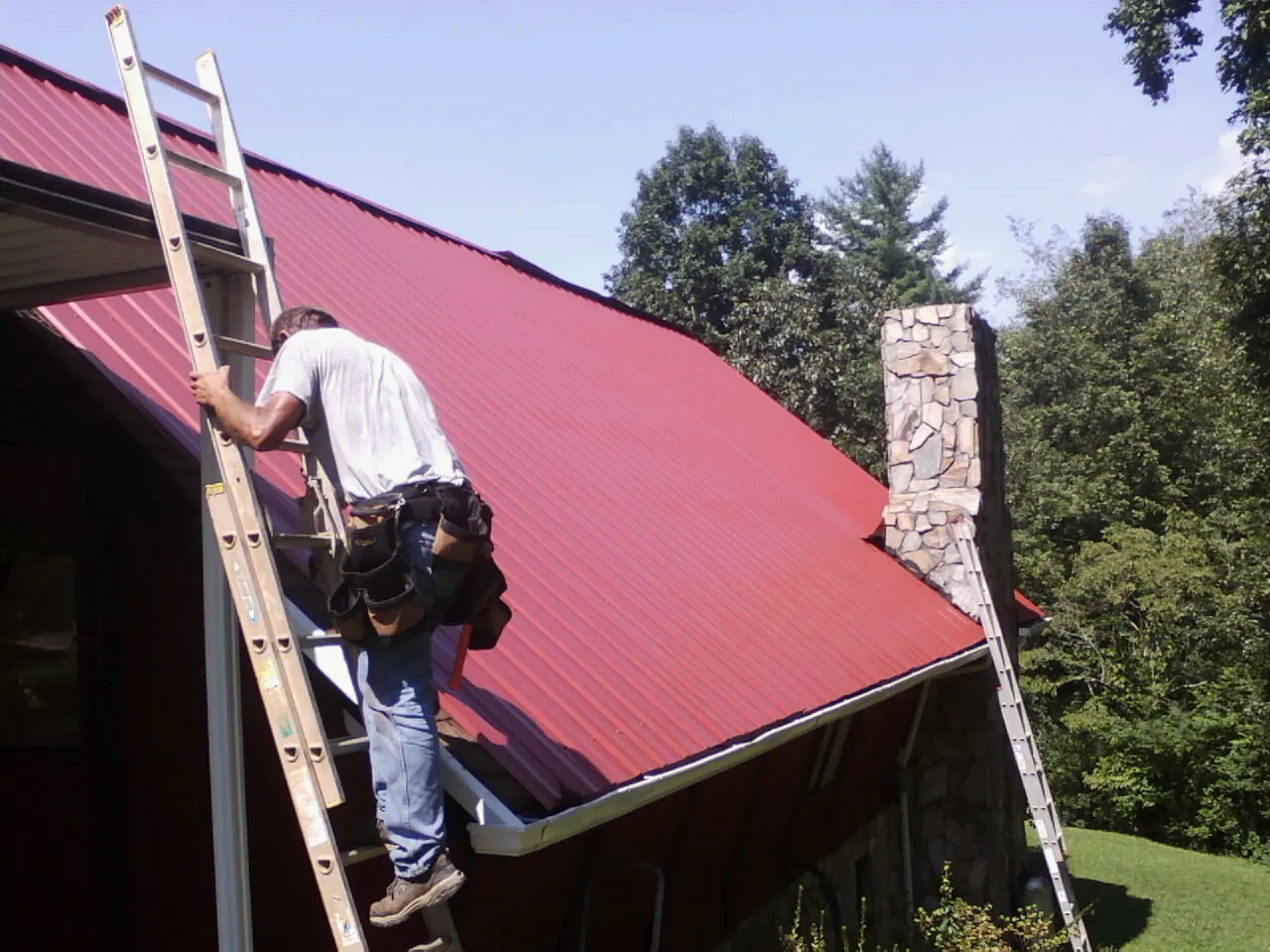 Roofer on a ladder beside a red metal roof and stone chimney — Soffit & Fascia Repair by Roofing Brothers in Eglon